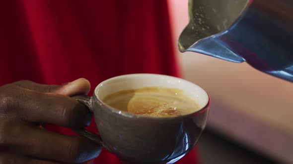 Midsection of african american barista pouring milk from jar into coffee cup in cafe