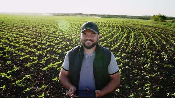 Happy Farmer Stands and Smile Holds Tablet in His Hands Against Background of Working Tractor in