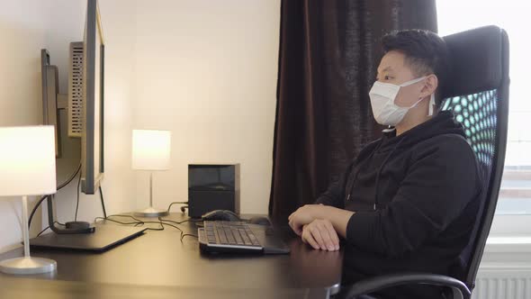 A Young Asian Man in a Face Mask Looks at the Desktop Computer Screen at Home Then Looks at Camera