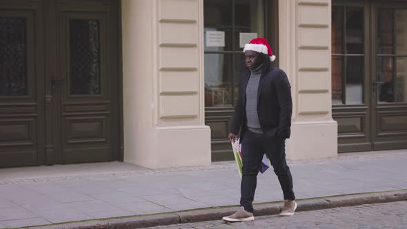 Black Man with Christmas Hat Walking on the City Street with Christmas Gifts