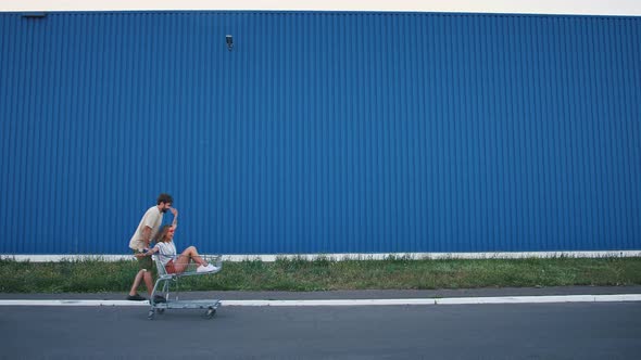Friends are Racing on Shopping Trolleys Against Blue Wall of Supermarket