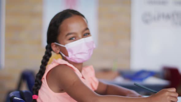 Portrait of mixed race schoolgirl wearing face mask, sitting in classroom looking at camera