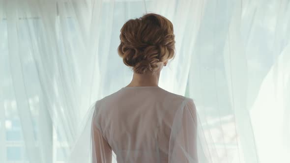 The back of the bride in a white dress opening the window. Wedding preparations