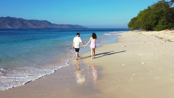 Young romantic couple holding hands while walking along the tropical white sand beach. Mountains in