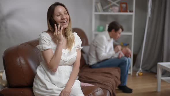Caucasian Woman Talking on Phone Laughing Sitting on Couch As Blurred Man Playing with Newborn Boy