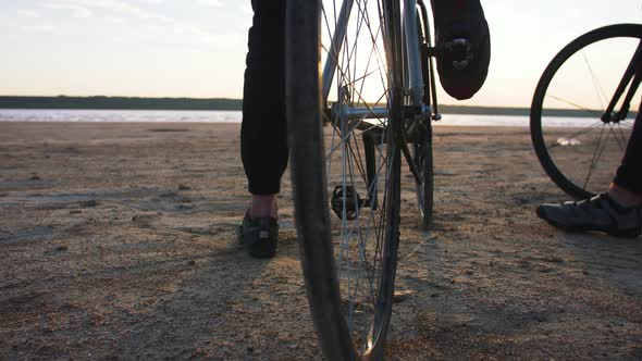 Two Young Men Sitting on Bicycles on the Beach on the Background of an Orange Sunsetting Sky Close