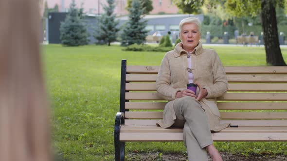 Relaxed Elderly Grandmother Drinking Coffee on Park Has Pleasant Conversation with Unrecognizable