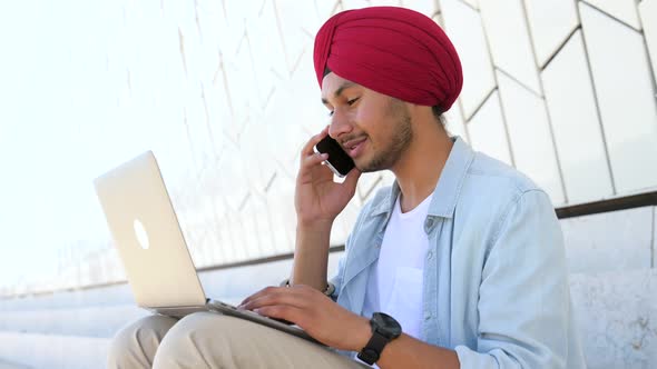 Cheerful Indian Male Entrepreneur with Laptop Talking on the Smartphone Making Schedule
