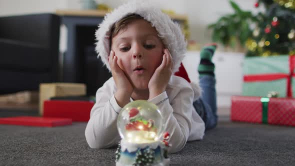 Surprised caucasian boy wearing santa hat lying on floor, watching snow globe