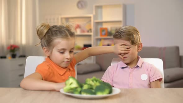 Little Sister Feeding Brother With Broccoli Covering His Eyes, Joking Friend