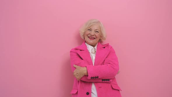 an Elderly Happy Woman in a Pink Coat Gestures with Her Thumbs Up While Posing in a Studio Against