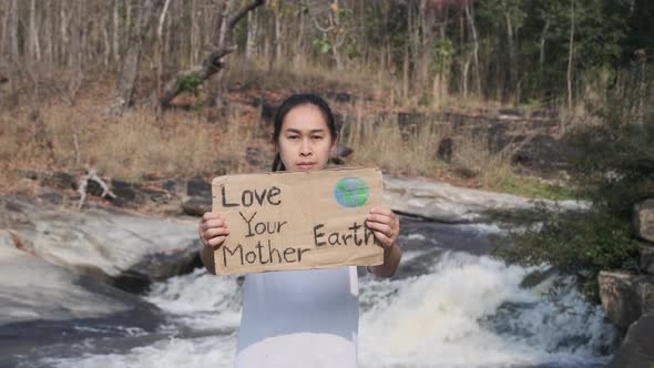 Portrait of woman standing with Love Your Mother Earth poster at a waterfall in the forest.