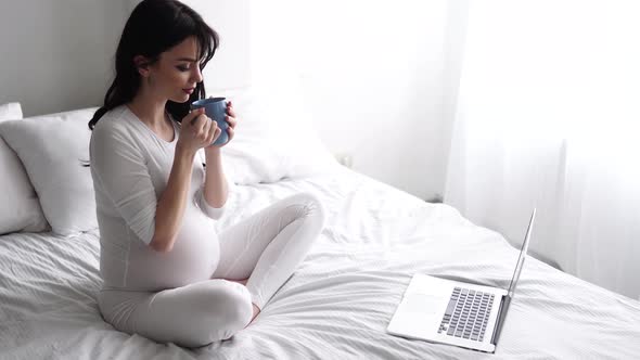 Pregnant Woman Drinking Tea, Using Computer At Home In Bedroom