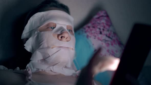 Young Woman in White Sheet Mask Lies on Blue Flowered Pillow