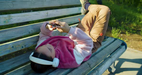 Ten Years Old Girl with Smartphone and in Headphones She Lying on the Bench and Chatting with