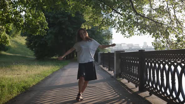 a Woman in Casual Clothes is Dancing Merrily in a City Park Near an Openwork Lattice