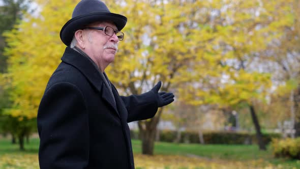 An Old Man Dressed in a Black Coat and Hat Tells the Story By Pointing His Hands to the Side