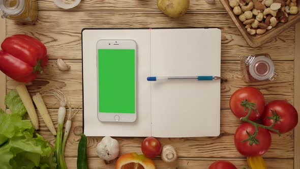 Pen Rolling on Blank Notebook with Smartphone on Kitchen Table with Vegetables
