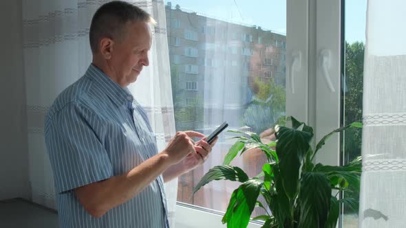 Elder Man Standing Near Window Indoors at Home While Using Mobile Phone