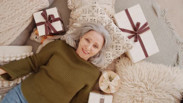 Cheerful Senior Woman Lying on Bed with Xmas Gifts and Posing for Camera