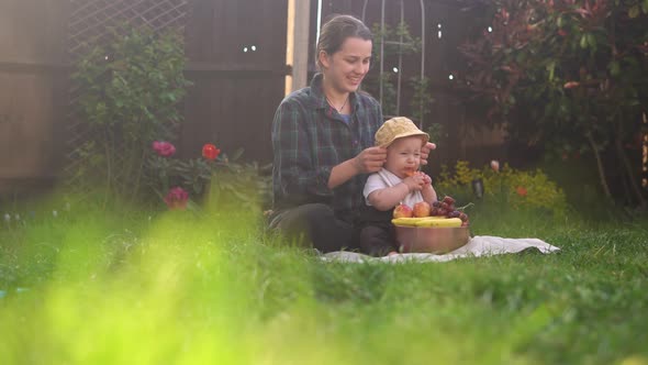 Happy Young Cheerful Mother Holding Baby Eating Fruits On Green Grass