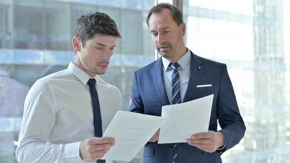 Two Businessmen Discussing Project on Documents Against Window