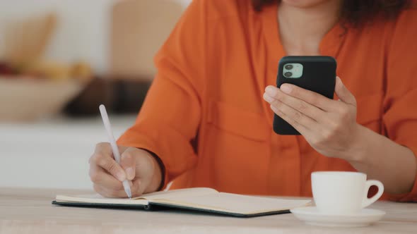 Unrecognizable African Woman in Orange Blouse Sits at Table at Home in Office in Cafe with Cup of