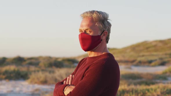 Portrait of happy caucasian man in face mask standing with arms crossed on sunny day at the beach