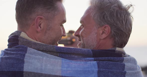 Happy caucasian gay male couple wearing blanket, embracing and taking selfie at sunset on the beach