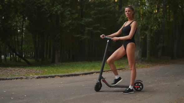 Confident Fit Woman Stands Near Her Electric Scooter and Looks at you at an Evening Park