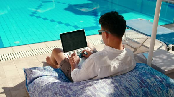 Man Works on Vacation Using Laptop By Swimming Pool in Hotel Resort in Summer