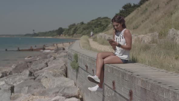 A Young Attractive Woman Sat By The Sea Using Her Phone To Order A Product Online - Ungraded