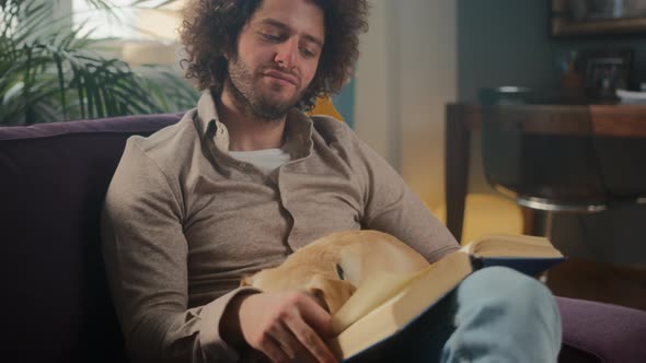 Handsome Curly Man Sitting at Home on the Couch with a Dog in His Arms and Reading a Book