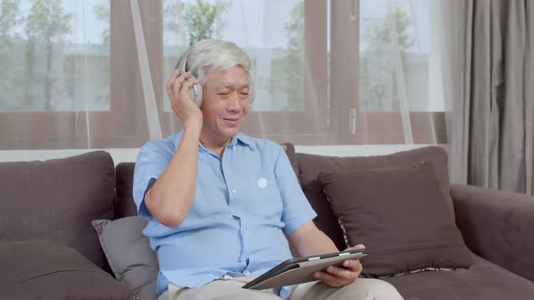 Asian older male happy wear headphone using tablet listening podcast while lying on sofa.