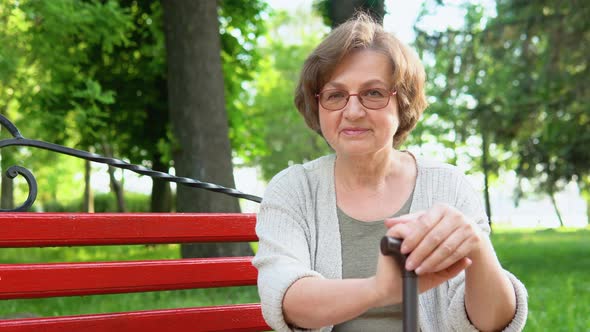 Portrait of Elderly Woman with Glasses and Cane Smiling on Bench in Public City Park