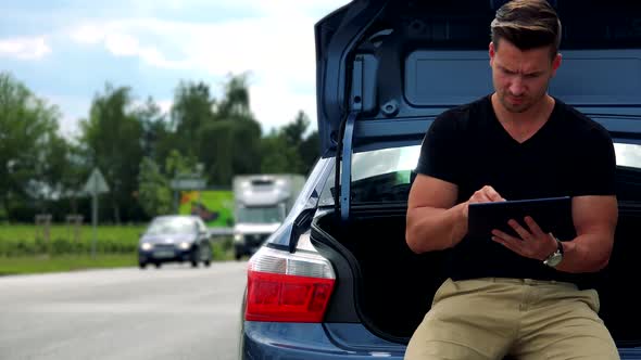 Young Handsome Man Sits in Car Trunk and Works (Typing) on Tablet-road with Cars