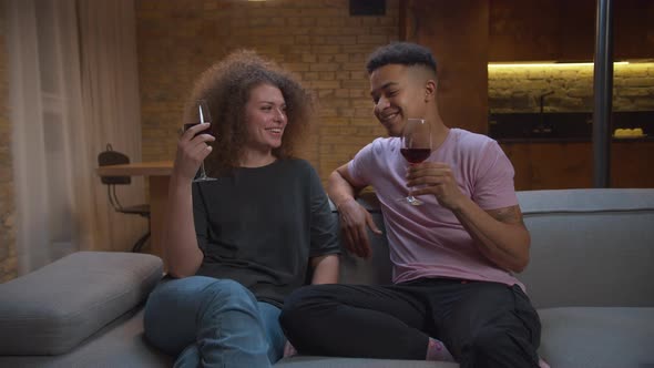 Young Mixed Race Couple Drinking Red Wine and Talking Sitting on Couch Together at Home