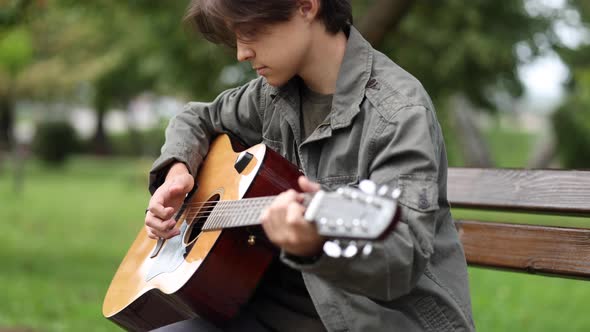 Teenage Boy Sitting on the Bench and Playing Guitar Outdoor in Park