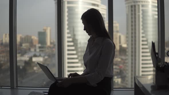 Young Woman Working on a Laptop Comfortably Sitting Near a Large Window