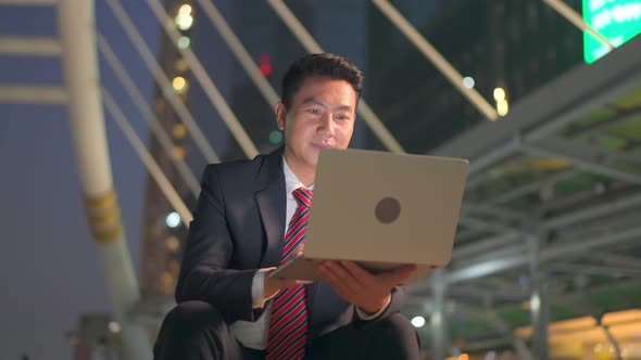 Asian businessman working on computer notebook sitting outdoor at night in city to complete job.
