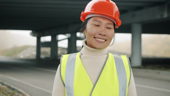 Asian Forewoman Wearing Vest and Helmet Standing Outside in Construction Site Smiling