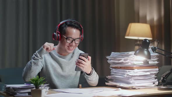 Asian Man With Headphones Listening To Music On Smartphone After Working With Documents At Home