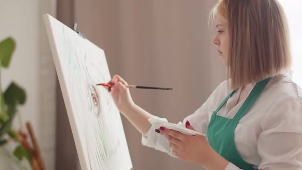 Young Woman Artist Paints on Canvas in Living Room Dipping Brush Into Palette Side View