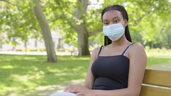 A Young Black Woman in a Face Mask Looks at the Camera and Sits on a Bench in a Park on a Sunny Day