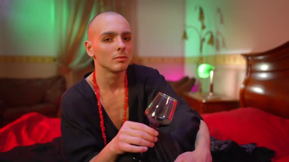 Depressed Male Queer Holding Wine Glass in Hand Thinking Sitting on Bed in Red Light