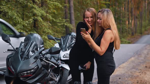 Smiling Women Friends Standing Near Their Motorbikes in the Forest and Looking at the Pictures They