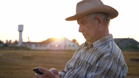 Elderly Man Farmer In Hat On A Rural Field At Sunset Uses A Smartphone