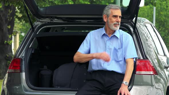 Senior Man Sits in the Trunk and Looks Around - Trees in the Background