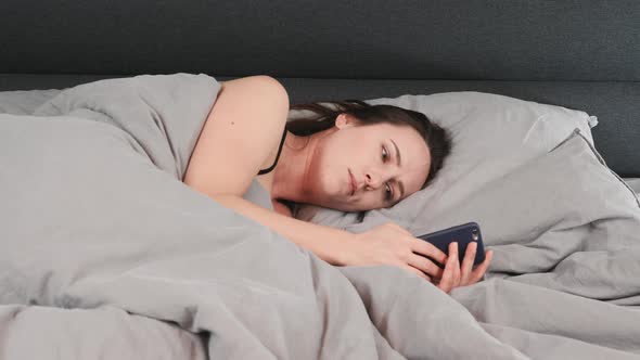 Portrait of young girl lying on grey bed and typing on smartphone. 