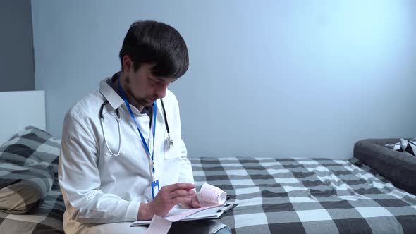 A Male Doctor Examines the Result of a Patients Electrocardiogram at Home Sitting on a Bed in the